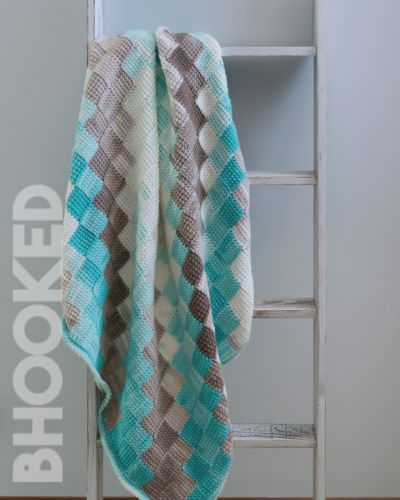 Baby Blanket crochet pattern diamond-pattern afghan in turquoise, brown, and cream draped over a white wooden ladder against a pale wall with a vertical watermark.